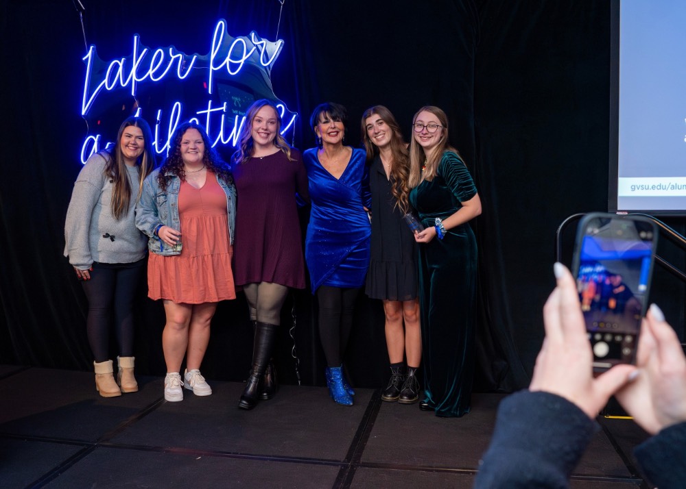 Group of 5 girls and President Mantella stand in front of Laker for a Lifetime neon sign with someone's hand in the corner taking the picture